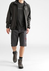 Mann, der einen schwarzen Outdoor-Jacke, ein schwarzes T-Shirt mit Logo, dunkelgraue Shorts mit Logo, Wanderschuhe und schwarze Socken trägt.