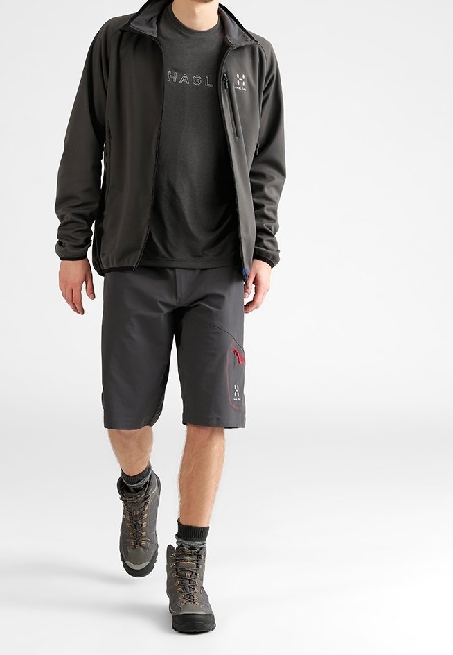 Mann, der einen schwarzen Outdoor-Jacke, ein schwarzes T-Shirt mit Logo, dunkelgraue Shorts mit Logo, Wanderschuhe und schwarze Socken trägt.