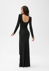 Kobieta w długiej czarnej sukience z niskim dekoltem na plecach i długimi rękawami, nosząca złote bransoletki, pozowała tyłem na jednolitym białym tle.
