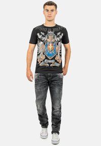 Zwarte t-shirt met grafisch ontwerp dat een blauwe schild, gouden accenten en adelaarmotieven toont, gecombineerd met grijze versleten jeans en witte sneakers.