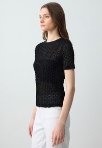 Top nero a maniche corte con un design trasparente e testurizzato caratterizzato da un motivo lavorato a maglia. Indossato con pantaloni bianchi, mette in risalto una lavorazione aperta.