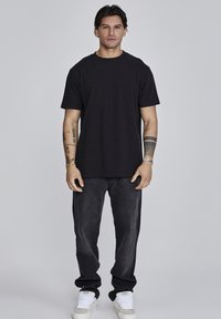 Camiseta de algodón negra con cuello redondo, combinada con jeans de mezclilla gris oscuro. El modelo lleva zapatillas blancas, mostrando un look casual.