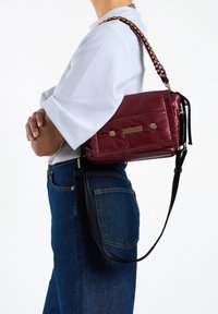 Bordeaux gekwilt schoudertas met gouden hardware, zwarte schouderband en geweven kleurrijke accenten, met een ritssluiting en een detail met voorvak.