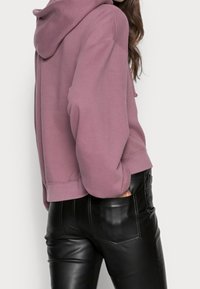 Sudadera recortada en tela de color rosa polvo con capucha ajustable, combinada con pantalones de cuero negros que tienen detalles brillantes y bolsillos frontales.