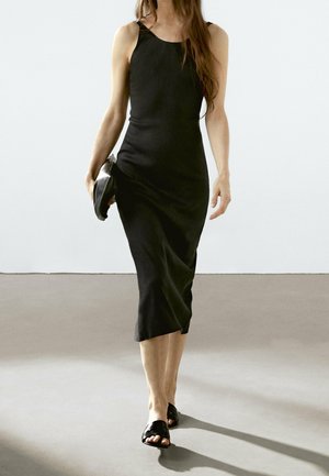 Woman walking in a black sleeveless midi dress, holding a black clutch in her left hand, wearing black open-toe slide sandals.