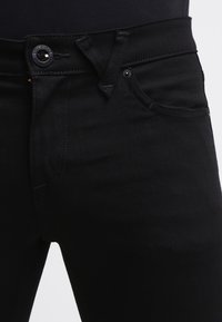 Jeans de mezclilla negra con corte ajustado, que cuentan con un cierre de botón, bolsillos frontales y detalles de costura en contraste. Textura suave y diseño minimalista.