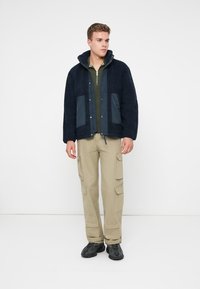 Mornarska flis jakna s kontrastnim slatko plavim džepovima, zatvaranjem na rajsferšlus i kapu, u kombinaciji s bež cargo hlačama i crnim tenisicama.