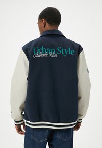 Giacca blu navy con maniche in pelle beige, con "Urban Style" e "Authentic Wear" ricamati in teal. Polsini e orlo a costine.