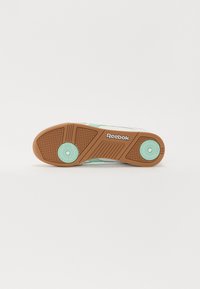 Soletenis Reebok s texturovanou hnědou gumovou základnou, mátově zelenými akcenty a kruhovým vzorem poblíž paty. Logo viditelné.