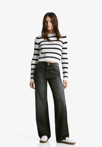 Maglione a righe in bianco e nero con costine, abbinato a jeans scuri a gamba larga a vita alta e sneakers bianche su uno sfondo neutro.