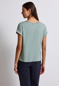 Top verde menta a maniche corte con una vestibilità rilassata, texture liscia, maniche arrotolate e scollatura a girocollo, abbinato a pantaloni scuri.