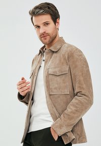 Tan suède jack met een knoopsluiting aan de voorkant, twee borstzakken en een puntige kraag. Gekozen over een wit topje en gestyled met textuurstof.