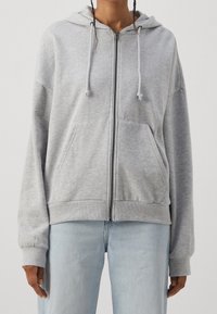 Šedá mikina na zip s kapucí se stahovací šňůrkou, předními kapsami a žebrovanými manžetami. Vyrobena z měkké látky s volným střihem a minimalistickým designem.