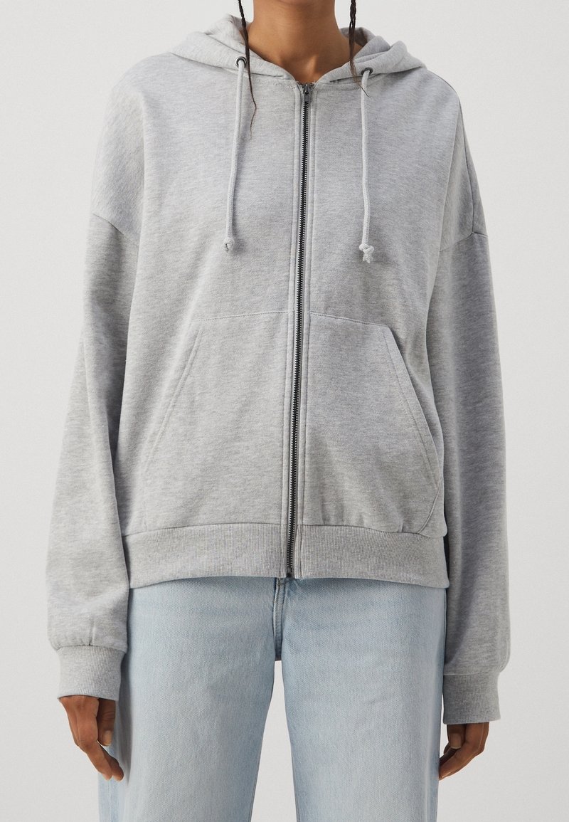 Sudadera gris con cremallera, capucha con cordón, bolsillos frontales y puños acanalados. Fabricada con una tela suave, de corte relajado y diseño minimalista.