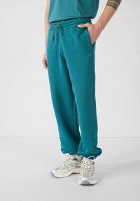 Teal sweatpants met een elastische tailleband, zijzakken en een relaxte pasvorm, gecombineerd met lichtgrijze sneakers met mesh-accenten.