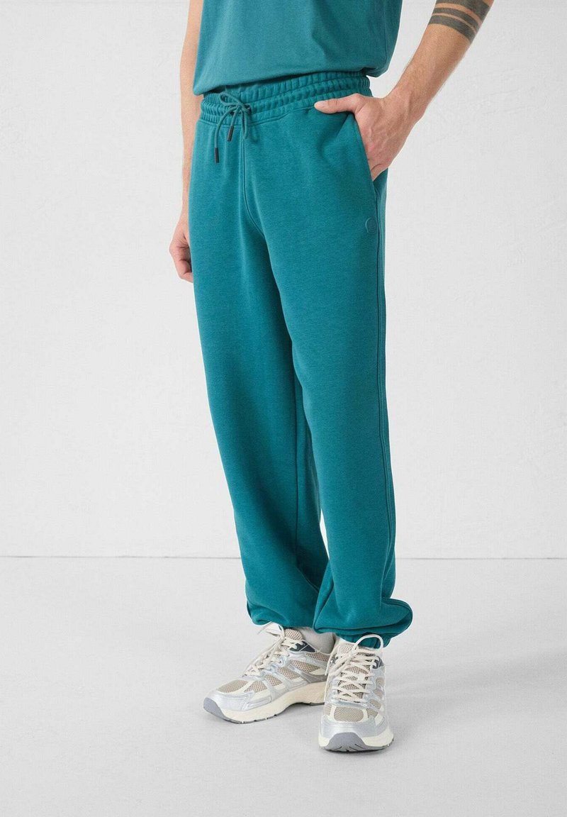 Teal sweatpants met een elastische tailleband, zijzakken en een relaxte pasvorm, gecombineerd met lichtgrijze sneakers met mesh-accenten.