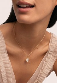 Collier en or avec un pendentif en forme de cœur orné d'une pierre blanche et de petits accents décoratifs, porté sur un haut léger à volants.