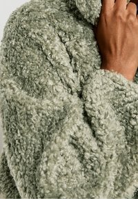 Chaqueta de gran tamaño, suave y de color verde claro, con un alto cuello y un tejido texturizado y aterciopelado. Las mangas tienen un ajuste relajado y llegan por debajo de la muñeca.