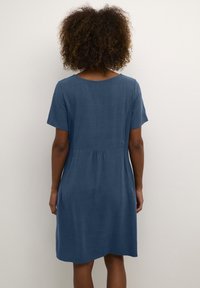 Short-sleeved, A-line dress in denim blue fabric. Features a round neckline, rear pleating, and side pockets. Smooth texture and casual design.