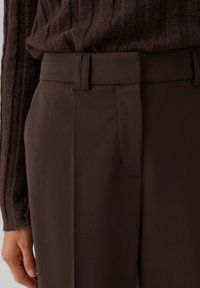 Close-up van bruine, hooggetailleerde, op maat gemaakte broek met ceintuurlussen, gedragen met een gestructureerde bruine trui en een ontspannen hand langs het lichaam.