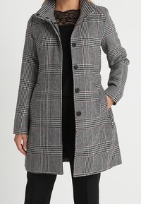 Cappotto scozzese in bianco e nero con sottili accenti rossi, dotato di colletto a scollo, bottoni frontali e tasche laterali. Finitura in tessuto textured.