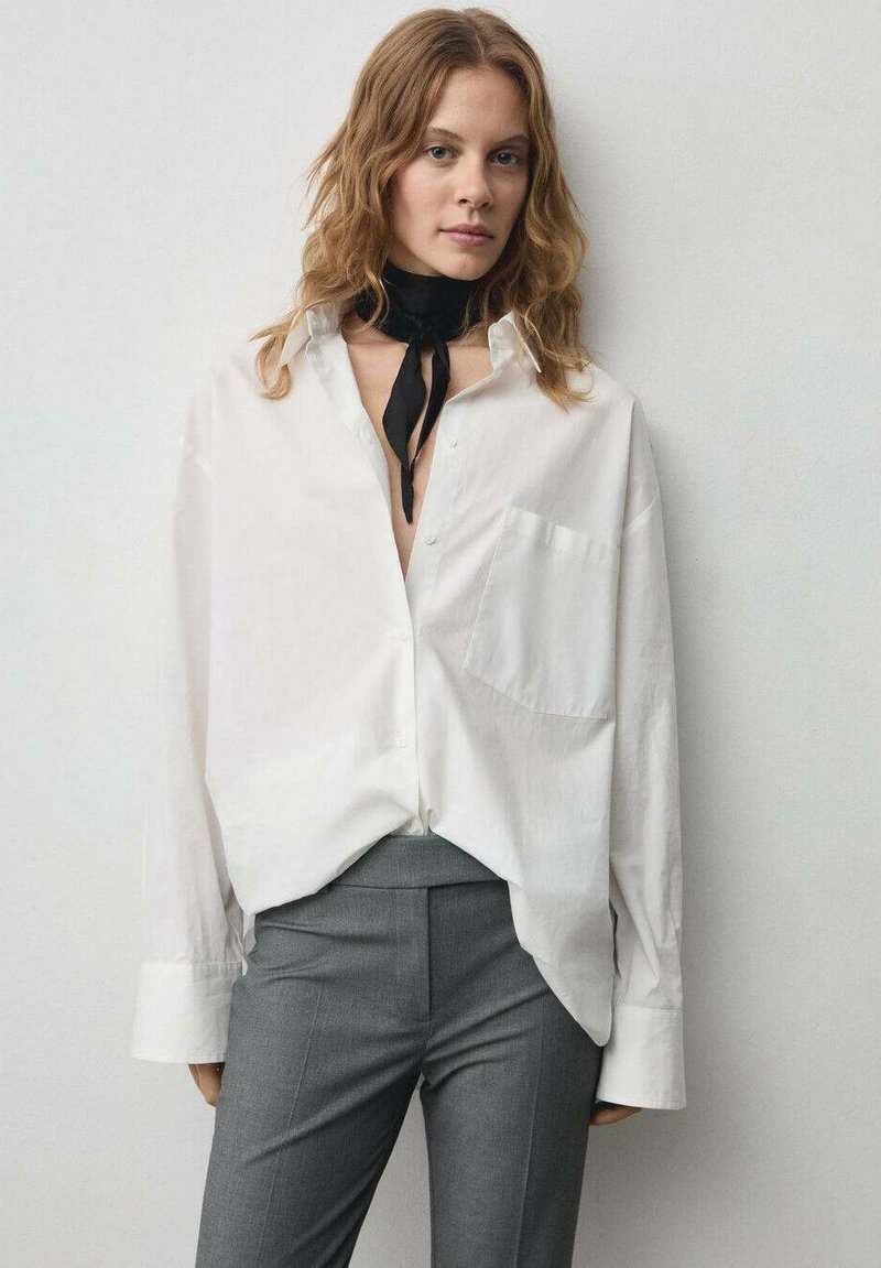 Bijela oversized košulja na dugme s džepom na prsima, uparena s krojenim sivim hlačama. Model nosi crnu kravatu, naglašavajući opušteni izgled.