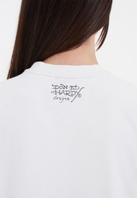 Witte katoen t-shirt met een ronde hals, met een zwart bedrukt ontwerp op de achterkant waarop "Don Ed Hardy designs" staat.