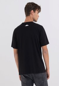 Camiseta de algodón negra con cuello redondo, mangas cortas y un pequeño logo blanco impreso en la parte posterior, combinada con pantalones vaqueros de mezclilla gris.