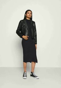 Svart läderkort jacka över en åtsittande svart klänning, ihop med svarta hög-top sneakers, vilket skapar en lager-på-lager urban look. Minimalistisk design.