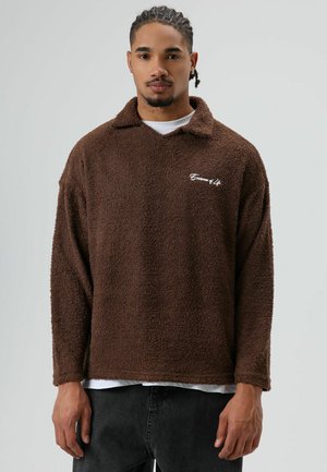 Bruine fleece trui met een losse kraag, met een zachte textuur en witte geborduurde tekst op de linkerborst. Gedragen over een wit overhemd.