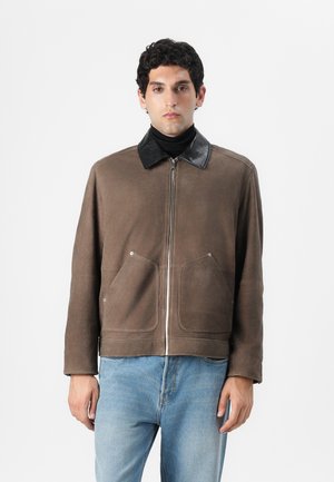 Brown leather jacket with a black faux leather collar, featuring two front pockets and a zip closure. Worn over a black turtleneck.