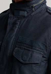 Marineblauwe utiliteitsjack met een hoge kraag, ritssluiting en twee opgezette zakken. Gemaakt van duurzaam stof met een gestructureerde afwerking en knoopdetails.