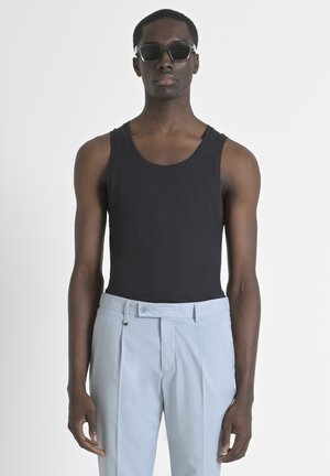 Débardeur noir à texture côtelée porté avec un pantalon plissé bleu clair. Le modèle porte des lunettes de soleil sombres. Design simple et épuré.