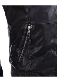 Schwarze Lederjacke mit glatter Textur, ausgestattet mit einem seitlichen Reißverschluss und geripptem Abschluss. Das Design umfasst detaillierte Nähte und kontrastierende Beschläge.