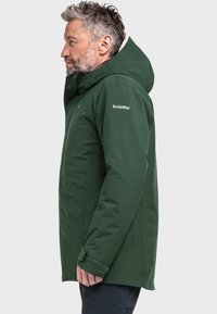 Donkergroene geïsoleerde jas met capuchon, voorzien van een gladde textuur, subtiele stikdetails en een logo op de rechter schouder.
