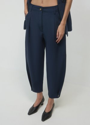 Marineblaue maßgeschneiderte Hosen mit Falten, umgeschlagenen Säumen und Knopfdetails. Glatter Stoff, weite Beine, kombiniert mit schwarzen spitzen High Heels.