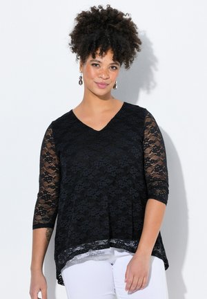 Top in pizzo nero con scollo a V e maniche 3/4; presenta una vestibilità fluida e un orlo scallopato. Indossato sopra pantaloni bianchi.