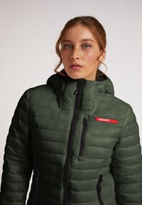 Grøn pufferjakke med hætte, der har vandret quiltning, sorte lynlåse og et rødt logo patch, der står "JACKIT."