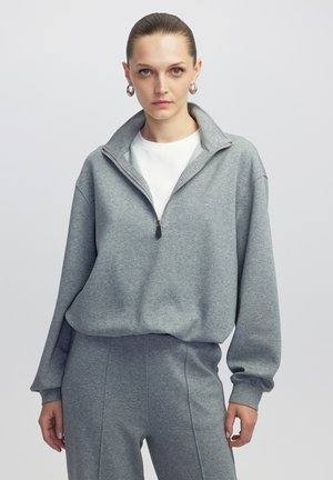 Sudadera gris con cremallera en el cuello, hecha de una tela suave, con un corte relajado, hombros caídos y puños acanalados para un toque de textura adicional.