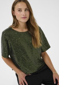 Vrouw met lang lichtbruin haar, gekleed in een donkergroene kortebloes met pailletten en zwarte broek, die iets opzij kijkt.