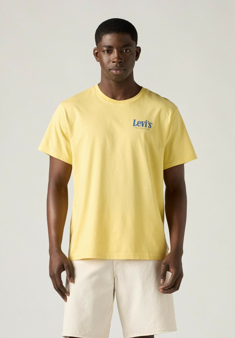 Junger Mann trägt ein gelbes Levi's T-Shirt und hellbeige Shorts und steht vor einem einfarbigen hellen Hintergrund.
