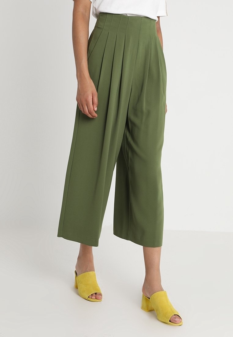 Nainen käyttää korkeavyötäröisiä oliivinvihreitä leveälahkeisia cropped-housuja ja keltaisia avokärkisiä paksupohjaisia mokkasiineja, seisten yksinkertaista taustaa vasten.