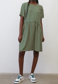 Vestido a cuadros verde y beige con mangas cortas, cintura fruncida y dobladillo a la altura de la rodilla. Combinado con zapatillas blancas con detalles en azul verdoso.