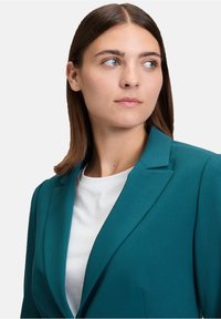 Teal blazer met een gestructureerd ontwerp, scherpe revers en een enkele knoopsluiting, gecombineerd met een wit T-shirt met ronde hals. Gladde textuur.