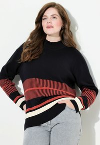 Schwarzer Strickpullover mit einem roten Streifenmuster im Brustbereich, cremefarbene Akzente an den Bündchen und einem hohen Halsausschnitt. Kombiniert mit grauen Jeans.