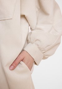 Cremefarbener Cord-Jackensaum mit gerillter Textur. Verfügt über ein umgeschlagenes Design und eine Seitentasche, die den weichen Stoff und die lockere Passform betont.