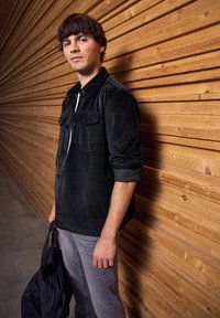 Černá kordurová košile s knoflíkovým zapínáním vpředu, dvěma náprsními kapsami a zahnuli rukávy, v kombinaci s světle šedými džíny a tmavou taškou. Dřevěné pozadí.