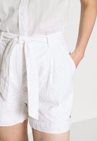 Hvide shorts med et struktureret blomstermønster, der har et bindebælte og sidelommer. Lavet af letvægtsstof, velegnet til varmt vejr.