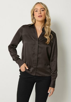 Blonde vrouw met golvend haar draagt een donkerbruine satijnen blouse met kraag en zwarte broek, staand met één hand in de zak.