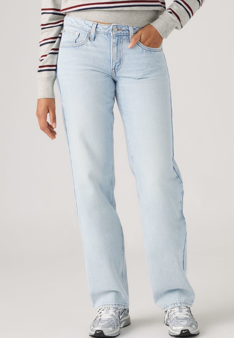 Personne portant un jean bleu clair droit, des baskets gris et blanc, et un haut rayé à manches longues avec une main dans la poche.
